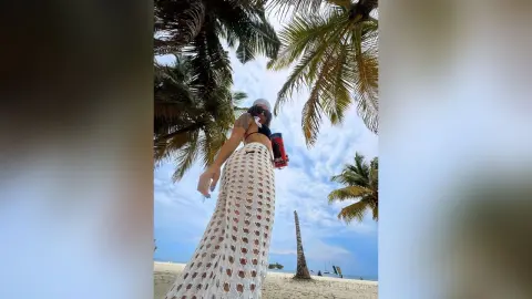AlesiaNoir — Woman in white crochet maxi skirt and bikini posing under palm trees on a sunny tropical beach.