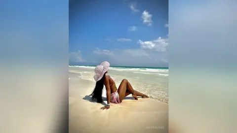 Woman in pink bikini and sun hat relaxing on a sunny tropical beach under a blue sky.