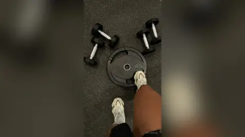 Person in white athletic socks sitting on a gym floor next to black dumbbells and a weight plate.