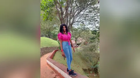 Smiling woman in a pink puff-sleeve top and blue jeans standing on a garden path near lush green trees.