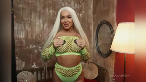Blonde woman in a lime green mesh two-piece outfit posing confidently in a stylishly lit room.