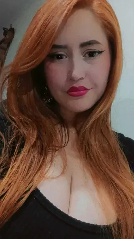 Redheaded woman in a black top with bold eyeliner and pink lipstick posing with a confident expression.