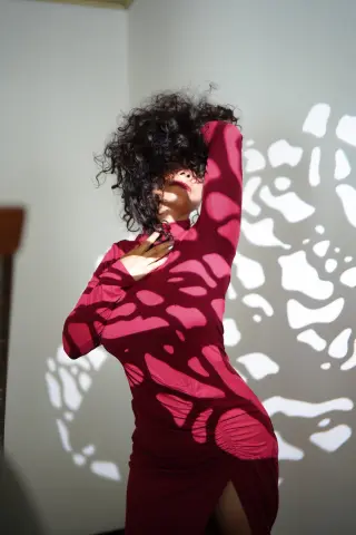 Woman in a long-sleeved burgundy dress standing against a wall with artistic leaf shadows.