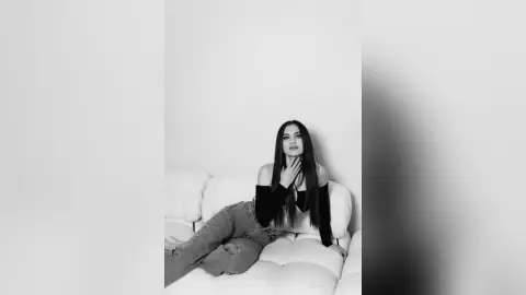 MilanaVolkova with long dark hair, wearing a black off-the-shoulder top and jeans, sitting on a white sofa against a plain white wall.
