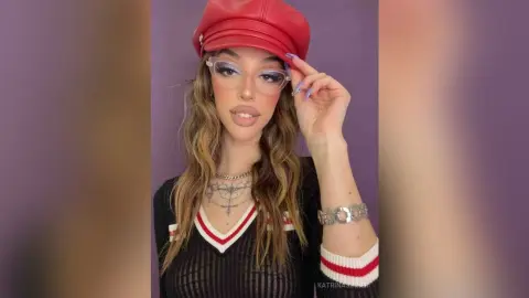 Stylish young woman in a red newsboy cap and black v-neck top posing against a purple background.
