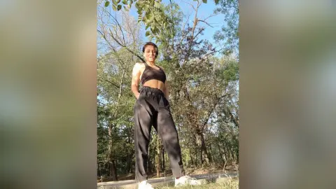 Athletic woman in black sports bra and joggers posing confidently in a sunny forest setting.