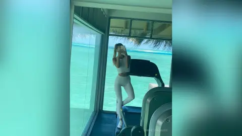 ChristineDavis poses in a white sports bra and leggings, taking a mirror selfie in a gym overlooking a bright turquoise ocean.