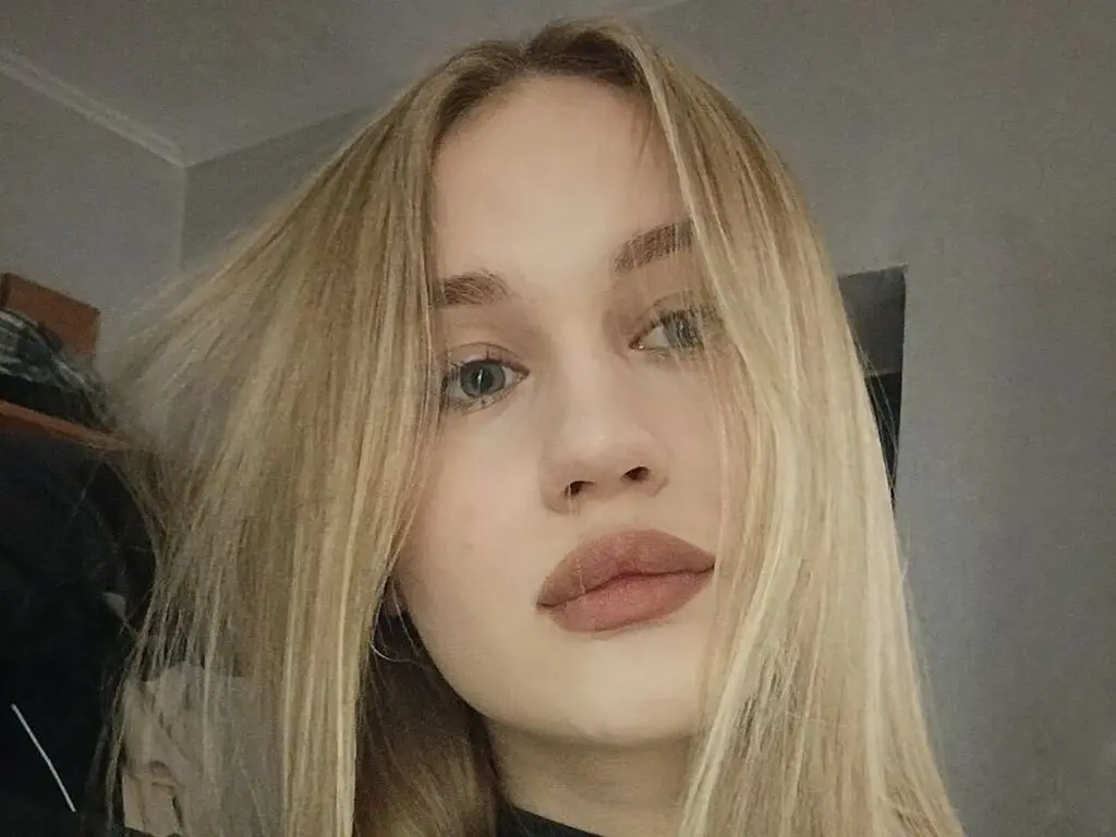 Close-up portrait of a blonde woman with natural makeup looking calmly at the camera in soft indoor lighting.