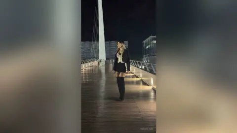 Stylish woman in a black mini dress and tall boots walking on a wooden pier at night under moonlight.
