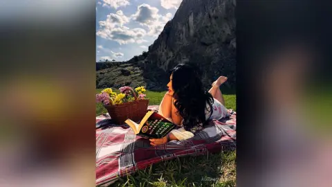 Woman in floral dress reading a book on a picnic blanket near a mountain under a sunny sky.