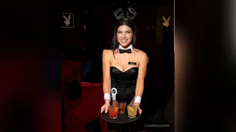 Smiling Playboy bunny hostess in black corset and bunny ears serving cocktails at a dark nightclub event.