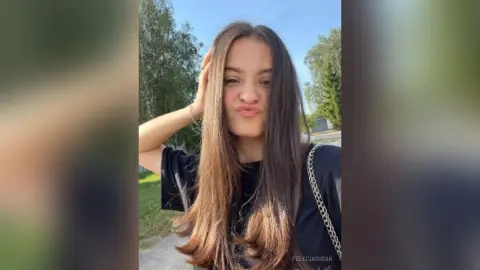 Young woman with long brown hair in a black t-shirt making a playful pouty face outdoors in sunny weather.