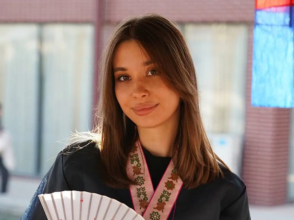 Smiling young woman in a black outfit and floral pink lanyard standing outdoors in soft daylight.