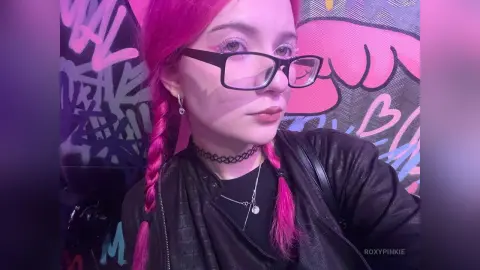 Young woman with pink hair and glasses wearing a black leather jacket against a colorful graffiti wall.