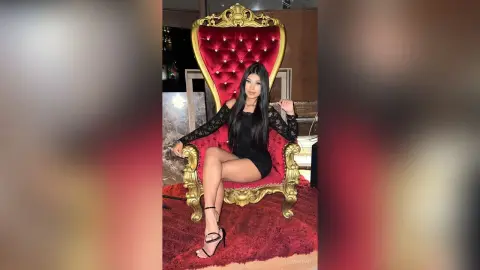 Elegant woman in a black lace dress sitting on a regal red velvet throne in a luxurious setting.