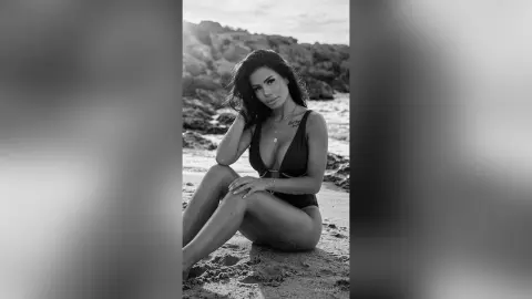 Moody black and white portrait of a woman in a dark one-piece swimsuit sitting on a sandy beach.