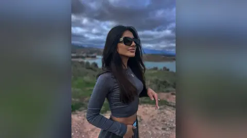 Stylish woman in a grey long-sleeve crop top and sunglasses posing against a scenic lake landscape at dusk.