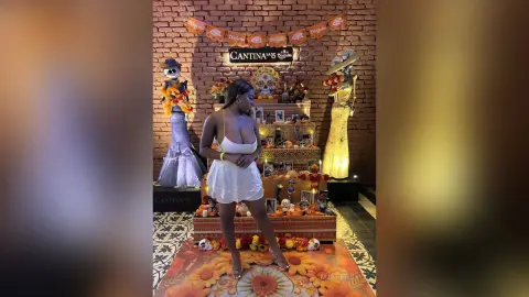 Woman in white dress standing by Dia de los Muertos decorations and skeleton statues in a festive cantina setting.