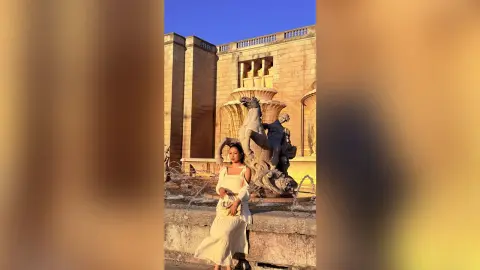 Elegant woman in a white summer dress standing by a classical stone fountain at golden hour.