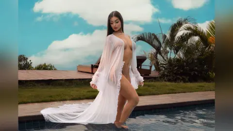 Elegant woman in a flowing white sheer robe posing by a luxury poolside under a bright blue sky.
