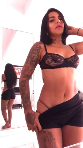 SusanaGlace poses in a black lace bra and black shorts, showing off her tattoos and midriff in a bright room.