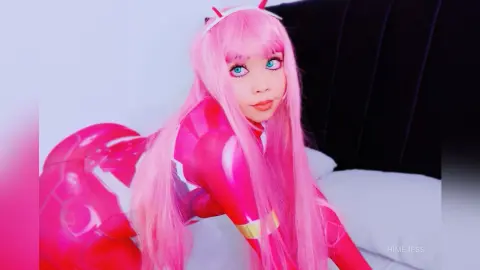 HimeJess poses in a pink bodysuit and long pink hair, looking at the camera while lying on a white bed against a dark headboard.