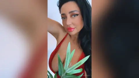 Glamorous woman with long dark hair in a red plunging dress posing with green leaves in a bright setting.