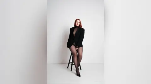 AryaSin poses sitting on a black stool wearing a black blazer over a sheer black bodysuit and black tights against a white background.