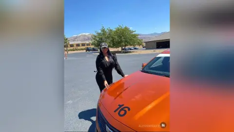 Stylish woman in black outfit standing next to an orange sports car in a sunny outdoor parking lot.