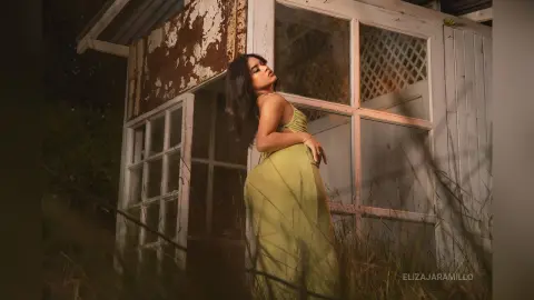 Elegant woman in a flowing lime green gown posing by a rustic white cottage at twilight.
