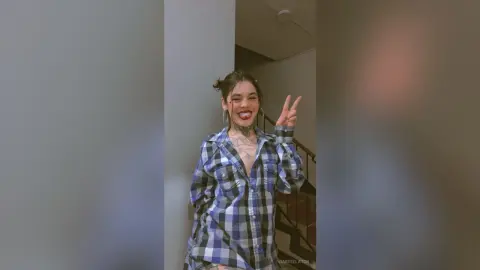Cheerful young woman in a blue plaid shirt making a peace sign in a modern indoor setting.