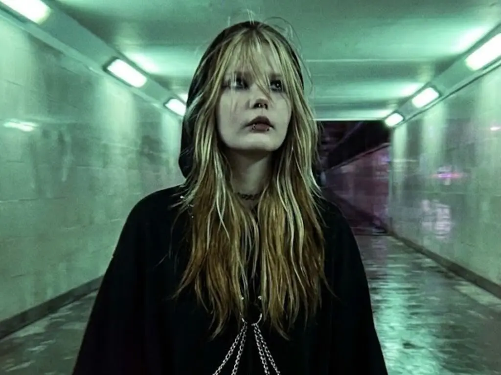 JaimieBez looks at the camera while holding a lit cigarette, wearing a black and tan jacket and a silver chain necklace.