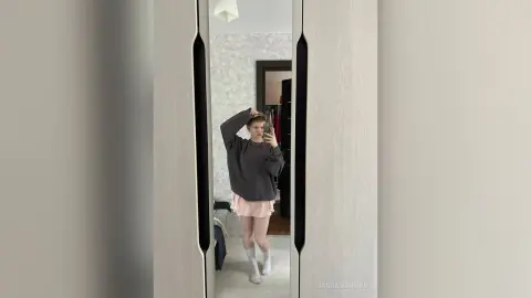Young woman in a grey sweatshirt and pink skirt taking a mirror selfie in a modern bedroom.
