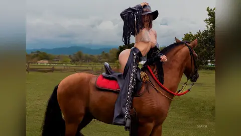 Woman in black leather cowgirl outfit riding a brown horse in a sunny green meadow.