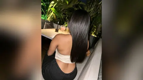 MiaKorz sits at an outdoor table with her back to the camera, wearing a white crop top and black pants.