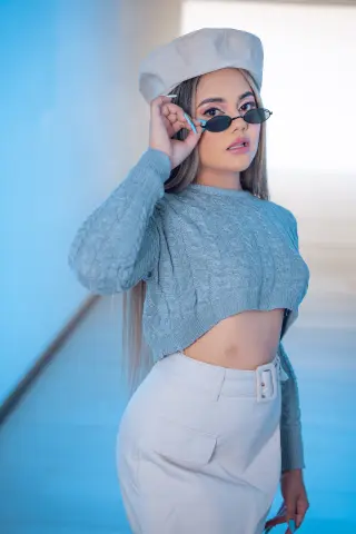 Stylish woman in a grey cable knit sweater, white beret, and sunglasses posing in a bright blue studio setting.