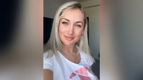 Smiling blonde woman in a white t-shirt posing for a bright, casual indoor selfie.