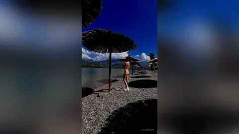 Slim woman in a bikini standing under a straw beach umbrella on a sunny, tranquil lakeside shore.
