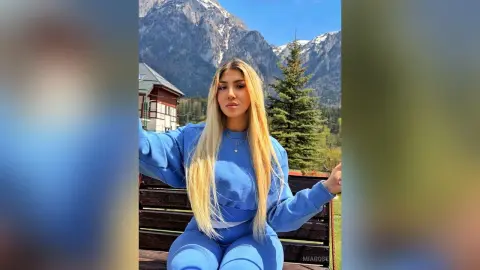 Blonde woman in a blue sweatshirt sitting on a wooden bench against a scenic mountain landscape.