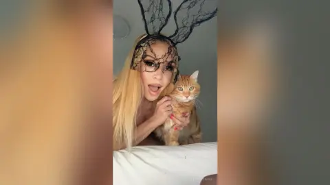 KendallMinx wearing a black lace spiderweb headpiece while holding an orange tabby cat.