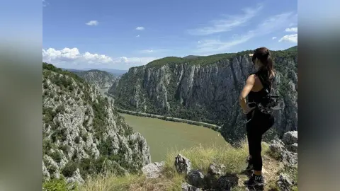 DyandraEve stands on a rocky cliff edge overlooking a deep river canyon under a clear blue sky.