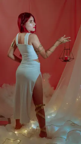 Red-haired woman in a white slit maxi dress holding a lantern against a dreamy pink cloud backdrop.