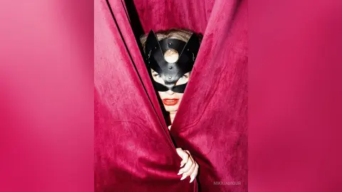 Mysterious woman in a black masquerade mask peeking through deep pink velvet curtains in a moody setting.