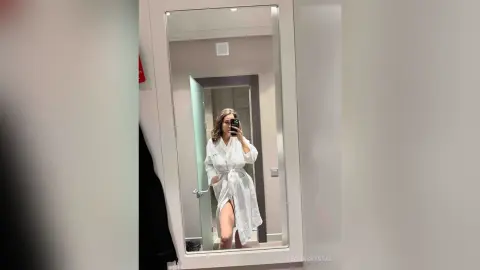 Young woman in a white textured robe taking a mirror selfie in a bright, modern bathroom.