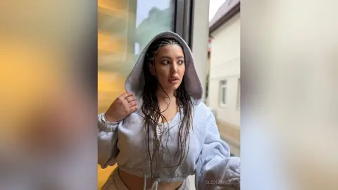 CleoLangley wears a grey hooded sweatshirt, looking off to the side with her long, dark braided hair visible under the hood.