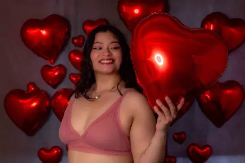 Smiling woman in a pink lace bralette holding a large red heart balloon against a dark background.