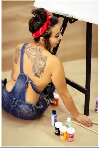 Woman in denim overalls sitting on a studio floor with paint jars, showing back tattoos in a creative mood.