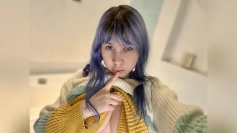 Young woman with blue hair wearing a cozy mustard yellow sweater in a bright, minimalist indoor setting.