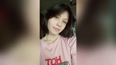 Young woman with dark wavy hair wearing a pink graphic t-shirt in a casual indoor setting.