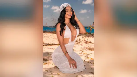 Smiling woman in a white lace beach outfit sitting on a sunny tropical sand beach.
